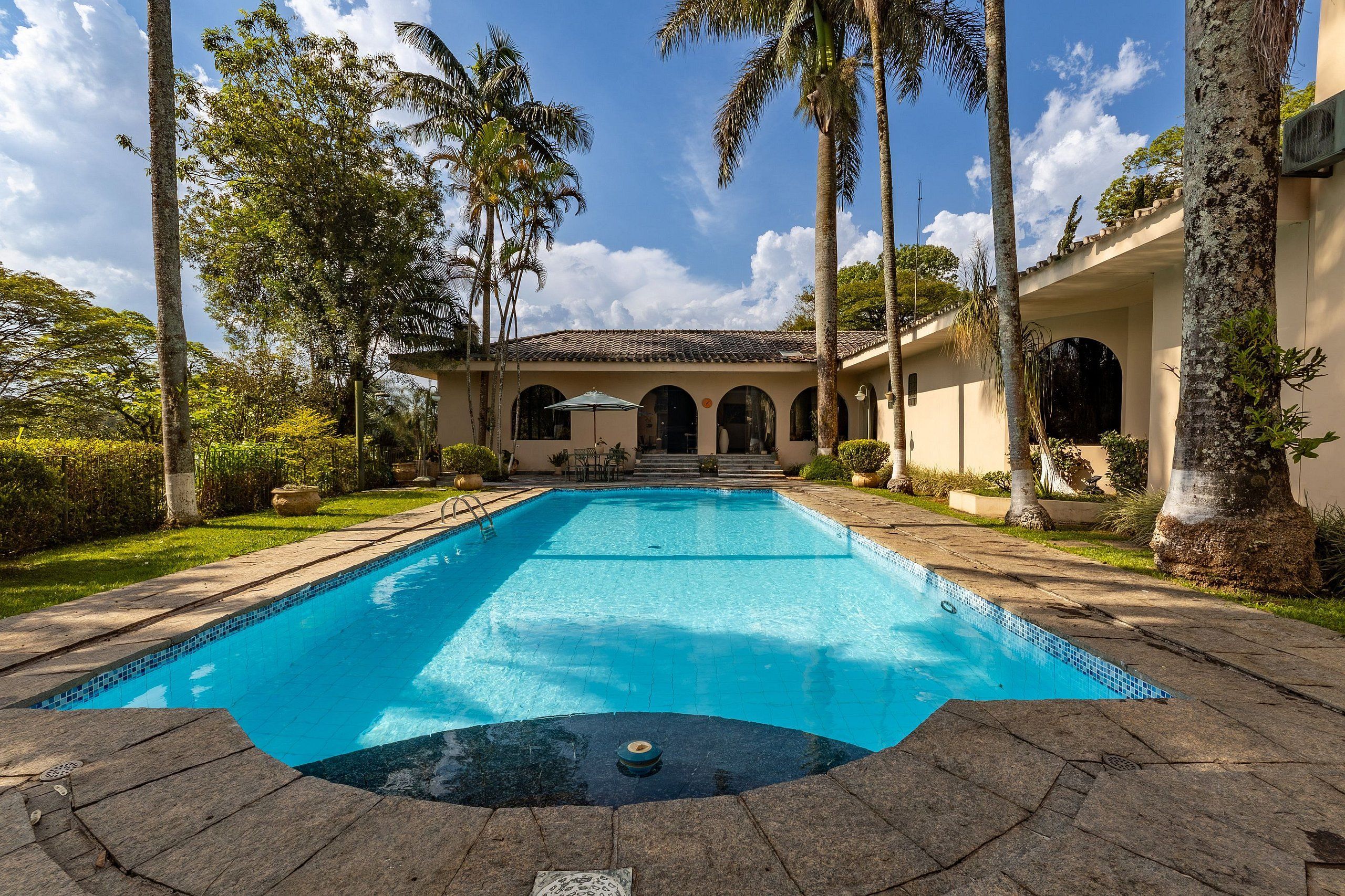 Ein Swimmingpool mit Fliesenrand, umgeben von Palmen und einem Gebäude mit Bogenfenstern.