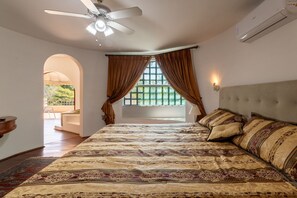 A bedroom with a large bed, a ceiling fan, an air conditioning unit, a window with curtains, and a view of greenery outside. - 5BR: Tennis Court, Sauna & Nightclub, 30min SP (Carapicuíba)