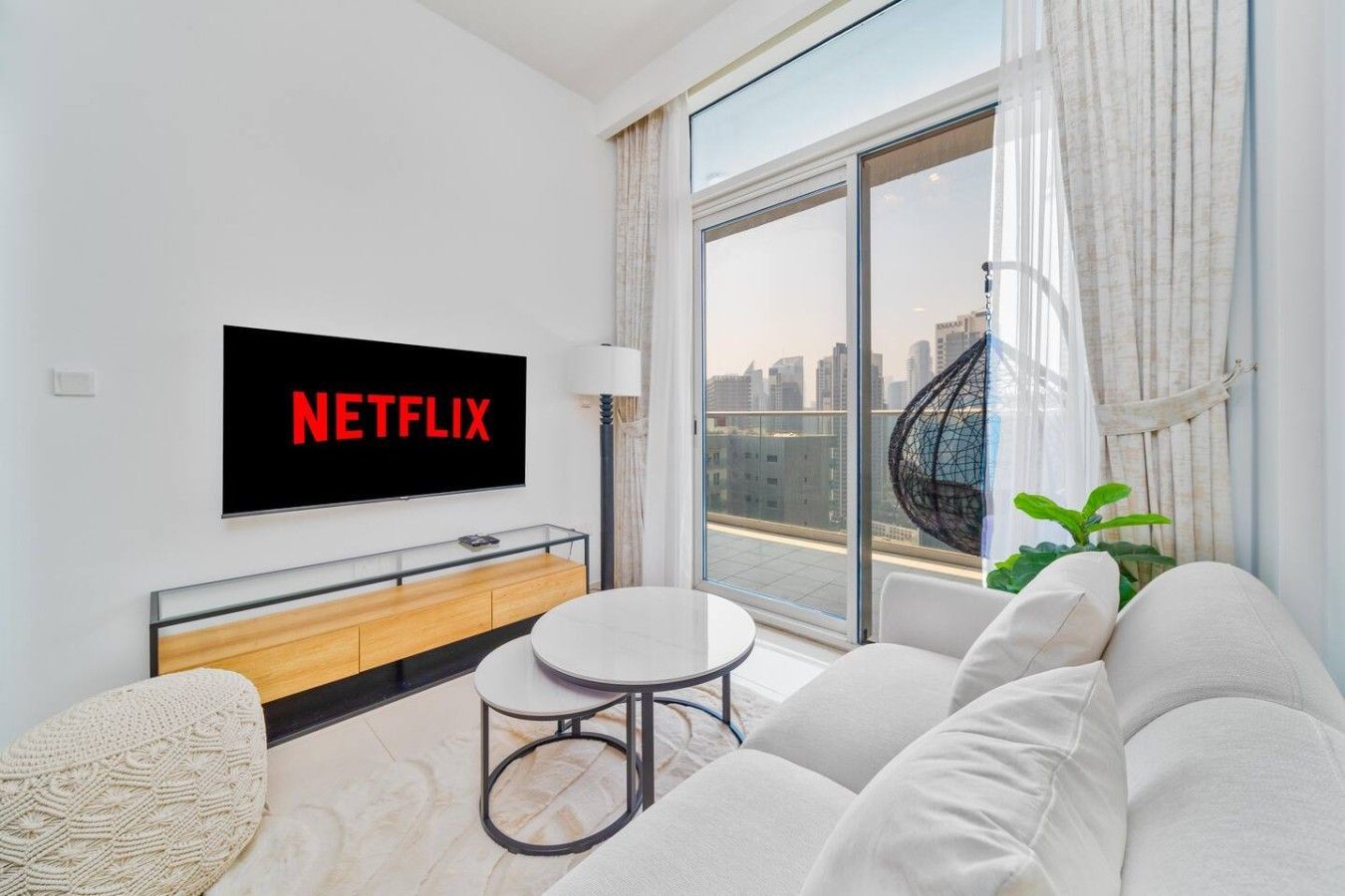 A modern living room with a large flat-screen TV displaying 'NETFLIX', a sofa, a small round table, and a view of a cityscape through large windows.