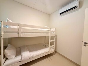 Ett rum med en våningssäng, inrett i vita nyanser, med en luftkonditioneringsanläggning monterad på väggen och en dörr på höger sida.