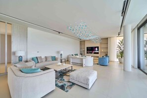 Een moderne woonkamer met een bank, salontafel, voetenbankje, een flatscreen-tv en een decoratieve plafondlamp.