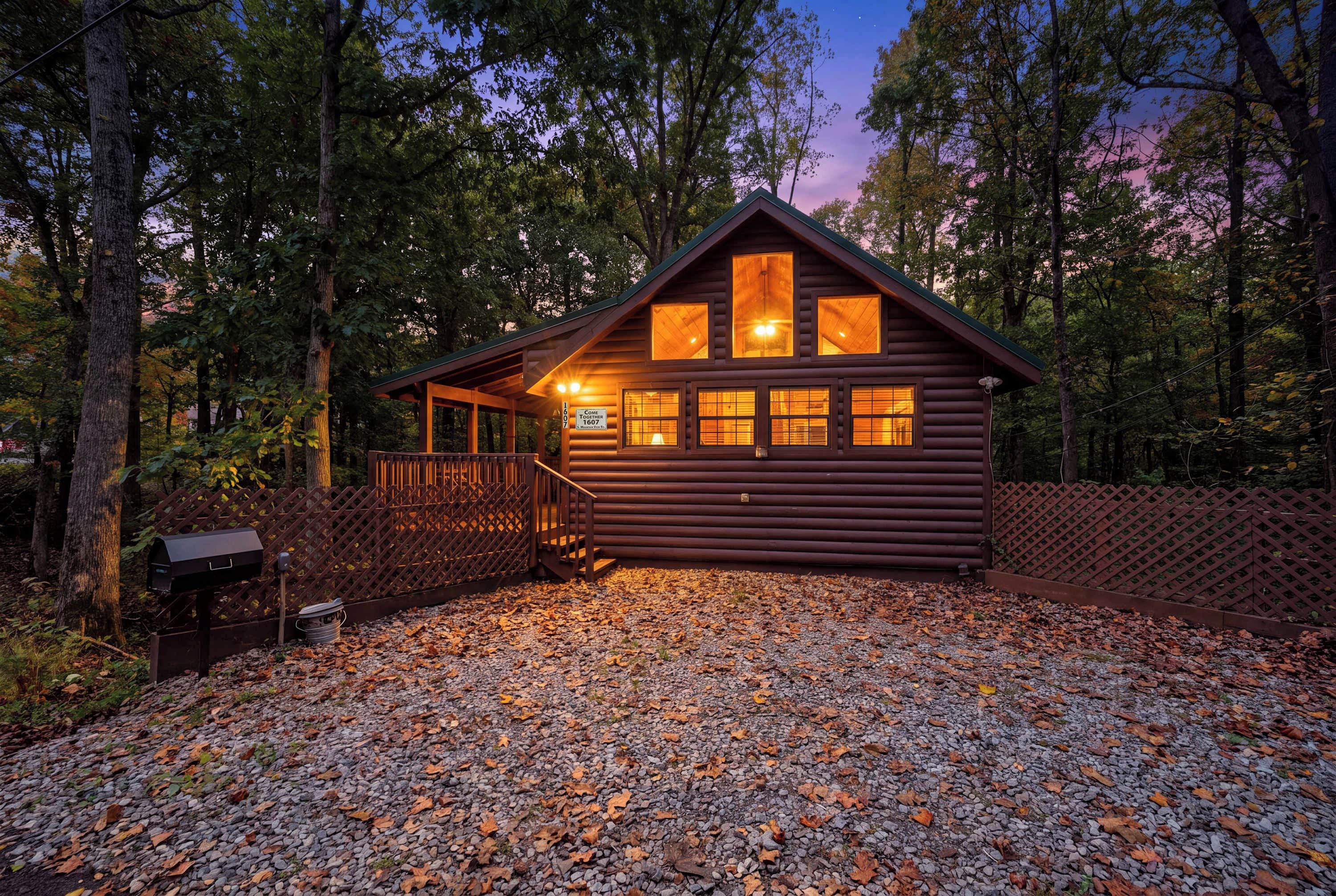 A wooden cabin with a porch, surrounded by trees and a gravel driveway.