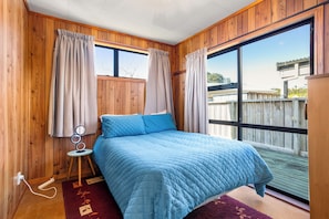 A bedroom with a bed, curtains, a small table with a lamp, and a view of the outside. - Kiwi Comfort - Whangamata Holiday Home (Whangamata)