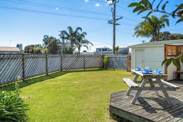 A backyard with a wooden deck, a picnic table, and a fenced area. - Kiwi Comfort - Whangamata Holiday Home (Whangamata)