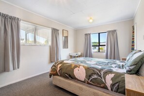 A bedroom with a bed, a window with curtains, a desk, and a view of the outdoors. - Kiwi Comfort - Whangamata Holiday Home (Whangamata)
