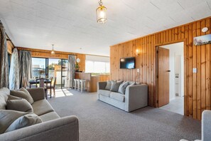 A spacious living area with a grey sofa, a wooden wall, and a television mounted on the wall. - Kiwi Comfort - Whangamata Holiday Home (Whangamata)
