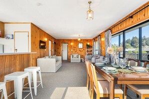 A room with a wooden interior, a dining area with a table set for four, and a view through large windows. - Kiwi Comfort - Whangamata Holiday Home (Whangamata)