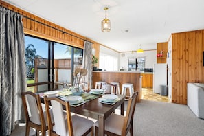 A dining area with a wooden table set for four, a kitchen in the background, and a view of the outdoors through large windows. - Kiwi Comfort - Whangamata Holiday Home (Whangamata)