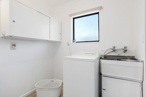 A compact laundry room with a washing machine, a dryer, a sink, and a window. - Kiwi Comfort - Whangamata Holiday Home (Whangamata)