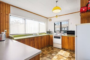 A kitchen with wooden cabinets, a white countertop, a stove, a sink, and a window. - Kiwi Comfort - Whangamata Holiday Home (Whangamata)