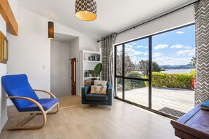 A spacious living room with a large sliding glass door leading to an outdoor deck, a blue armchair, a dark sofa with decorative pillows, and a bookshelf. - Harbour House - Whangamata Holiday Home (Whangamata)