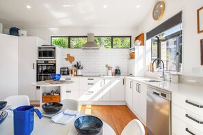 A modern kitchen with white cabinetry, a built-in oven, microwave, and dishwasher. There is a large window providing natural light and a view of greenery outside. - Harbour House - Whangamata Holiday Home (Whangamata)