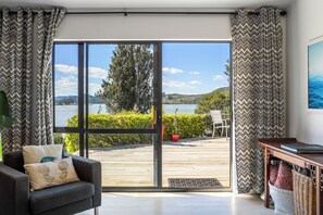 A modern living room with a large sliding glass door leading to a deck overlooking a lake. - Harbour House - Whangamata Holiday Home (Whangamata)