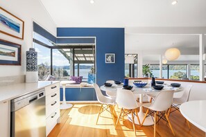 A modern kitchen with a view of the ocean, white cabinets, and a dining area with white chairs. - Harbour House - Whangamata Holiday Home (Whangamata)