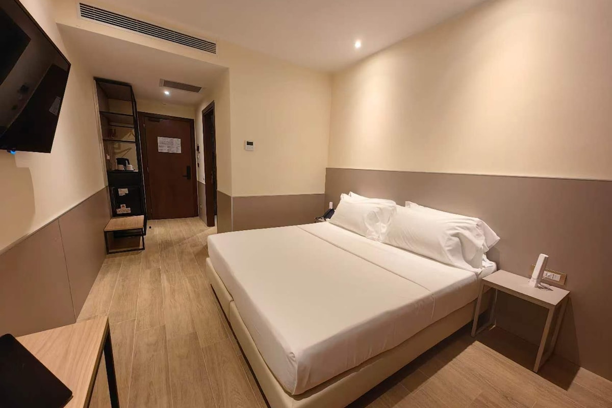 Una habitación de hotel con una cama grande, una mesita de noche, un televisor en la pared y piso de madera.