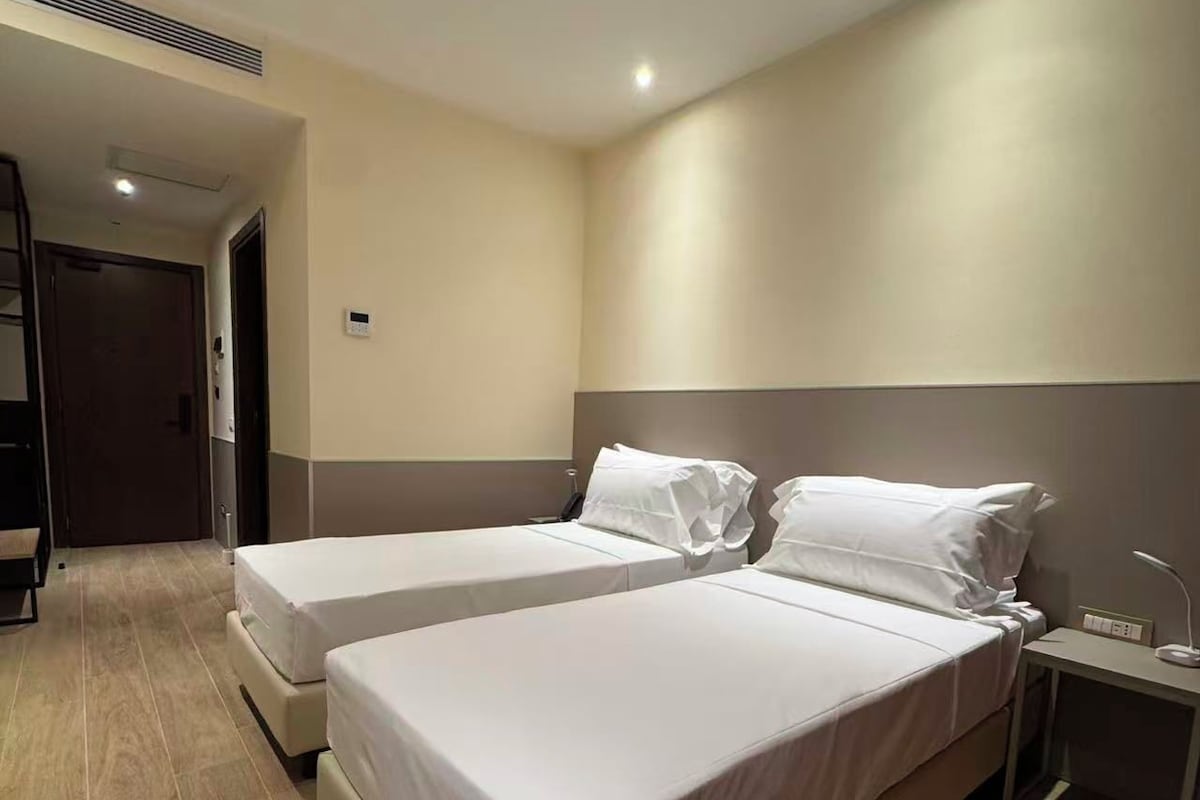 Habitación de hotel con dos camas, una mesita de noche y una puerta que da a un pasillo.