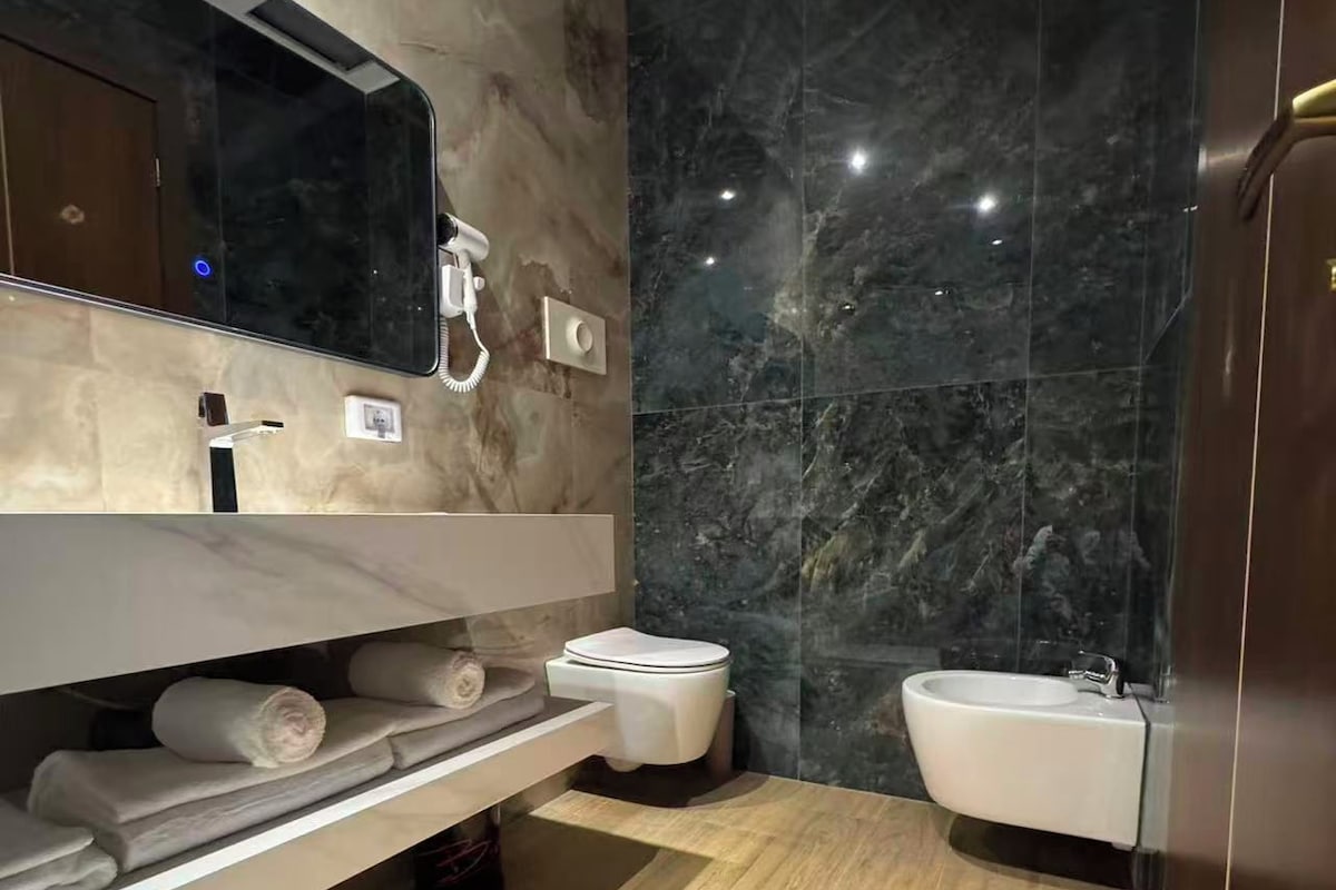 Un baño moderno con pared de mármol, tocador de madera, inodoro, bidé y lavamanos.