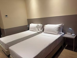 Habitación de hotel con dos camas individuales, una mesita de noche con lámpara y un termostato fijado a la pared.