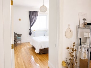 A bedroom with a wooden floor, a bed with white bedding, a chair, a window with curtains, and a wall-mounted shelf with items. - Pet-Friendly Farmhouse | Weddings, Special Events, Corporate Retreats + More! (Belleville)
