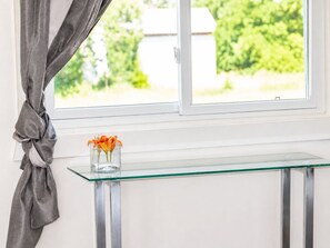 A window with a view of greenery, a glass table with a vase of flowers, and a curtain tied back.