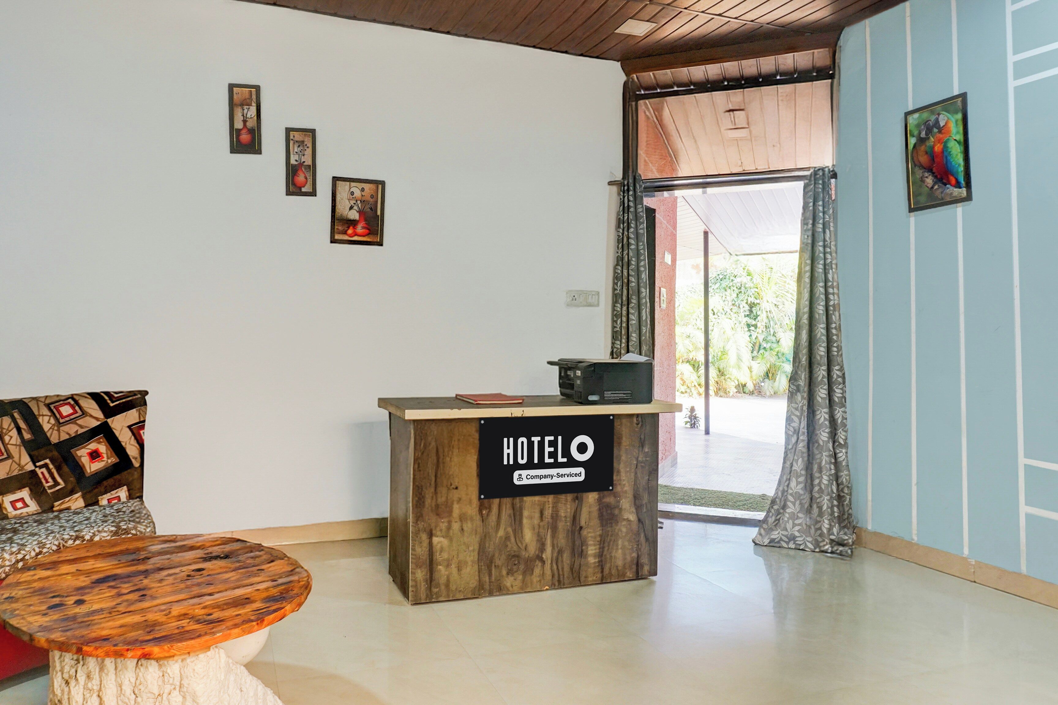 Zona de recepción con un escritorio de madera que dice 'HOTEL', un sofá con cojines estampados y vista al exterior a través de una puerta.
