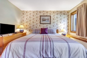 A hotel room with a large bed, two bedside tables, a flat-screen TV, floral wallpaper, and a window with curtains. - Kettlebrook | True Ski-In/Out | 3BR + Fireplace (Ludlow)