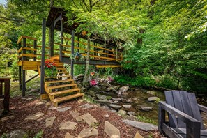 Uma ponte de madeira sobre um riacho, com uma escadaria de madeira que a conecta, cercada por vegetação exuberante.