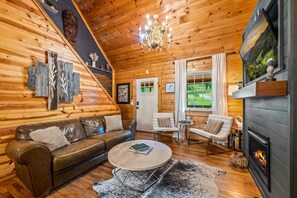 Ein gemütliches Blockhaus-Interieur mit Holzvertäfelung, einem Ledersofa, einem runden Couchtisch, einem Kamin und einem großen Fenster.