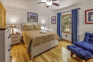 A bedroom with a wooden floor, a bed with a beige bedspread, a nightstand with a lamp, a chair, and a window with blue curtains. - Whispering Pines Retreat - Fire Pit - 2BR 2BTH (Rutherfordton)