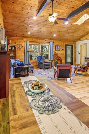 A cozy living room with wooden walls and ceiling, a dining table with a decorative runner, and a ceiling fan. - Whispering Pines Retreat - Fire Pit - 2BR 2BTH (Rutherfordton)