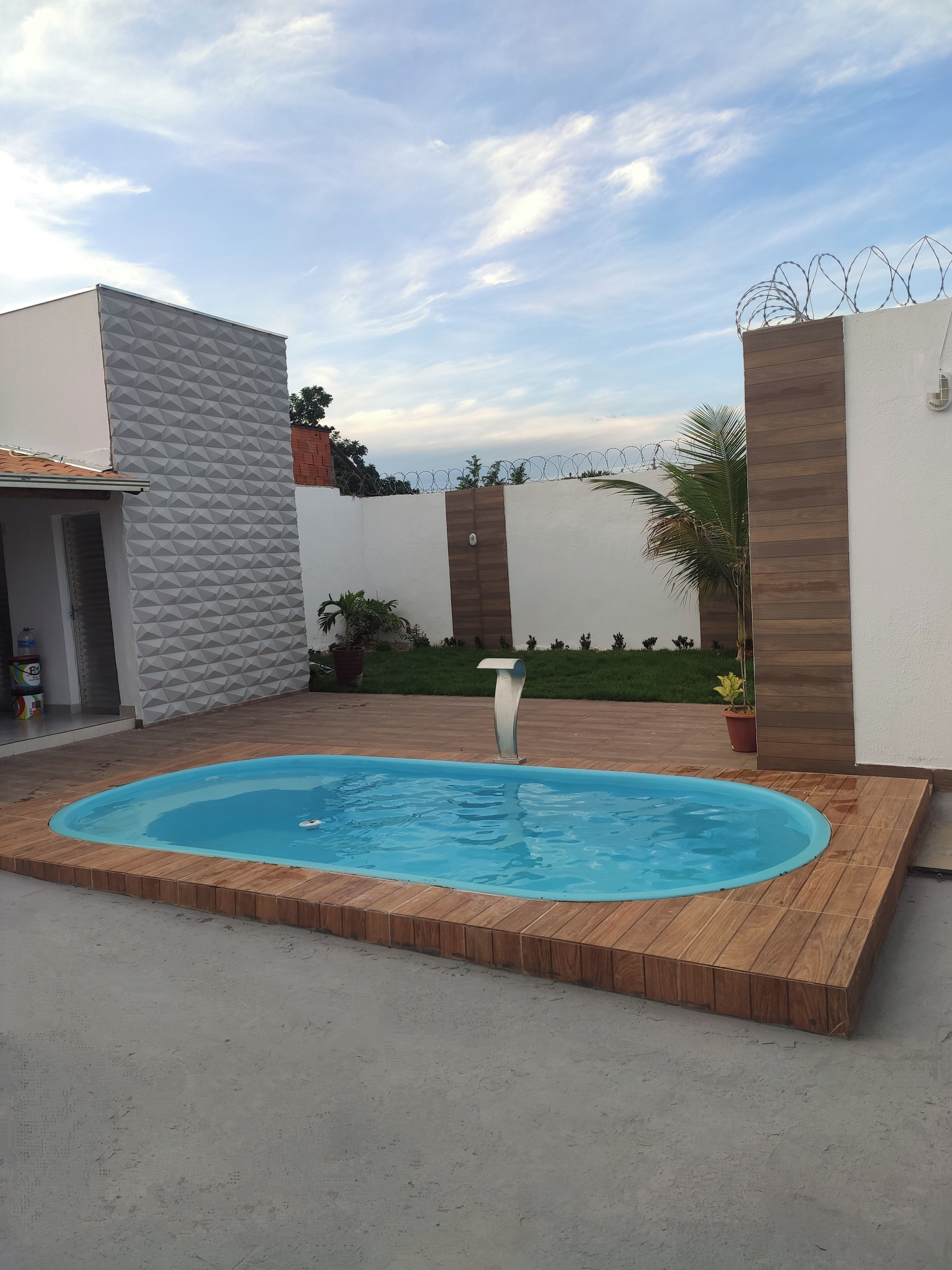 A swimming pool with a wooden deck, surrounded by a white wall with a geometric pattern, and a barbed wire fence.