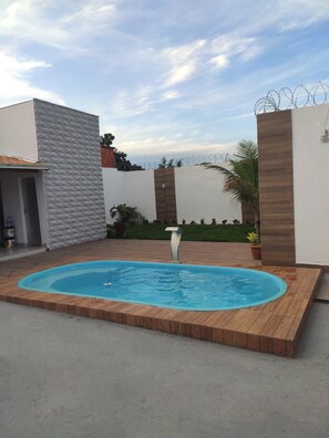 En swimmingpool med træterrasse, omgivet af en hvid mur med geometrisk mønster, samt et pigtrådshegn.