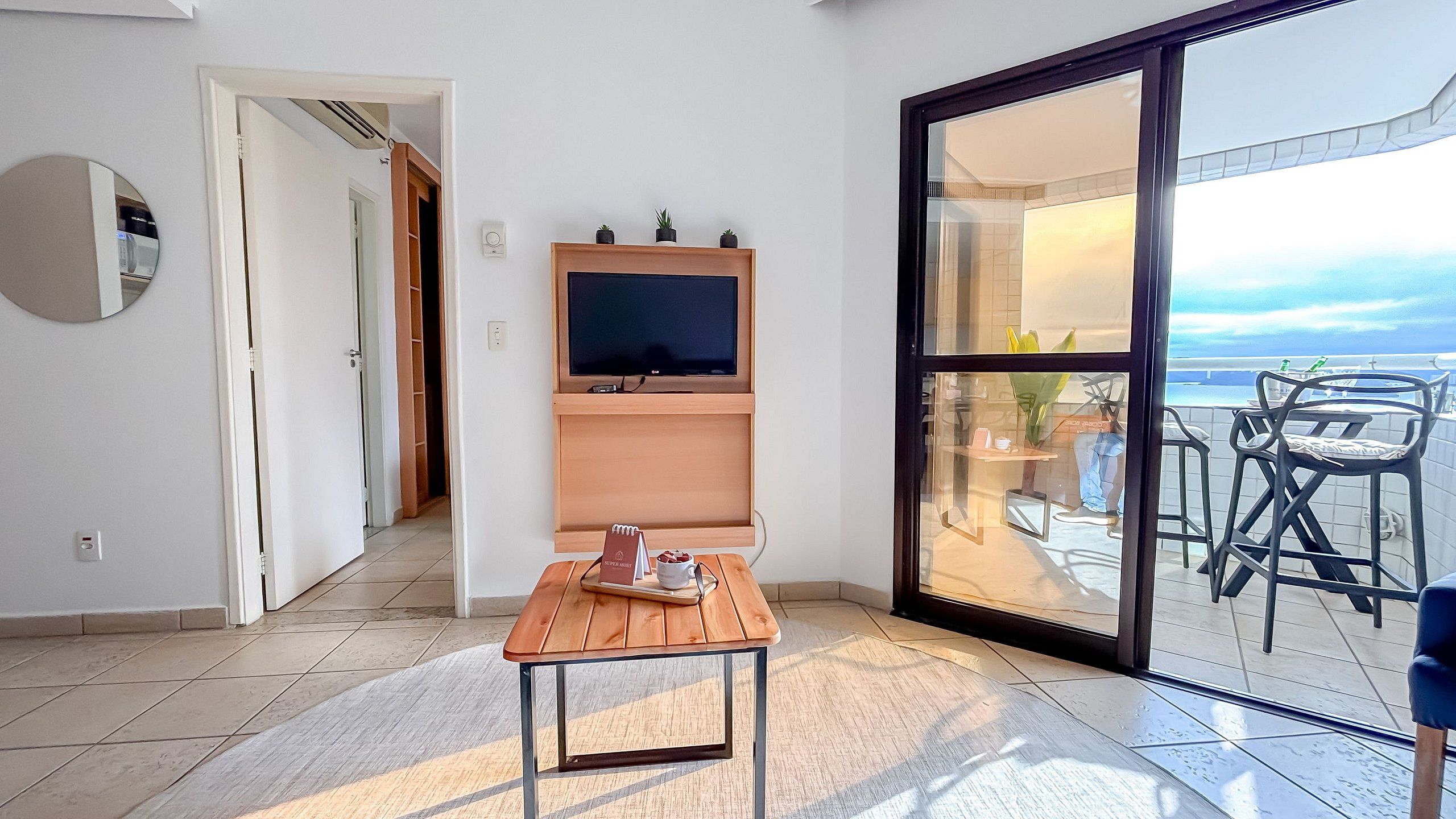 A room with a TV on a wooden stand, a small table with a notebook and mug, and a sliding glass door leading to an outdoor seating area with chairs and a view of the sea.