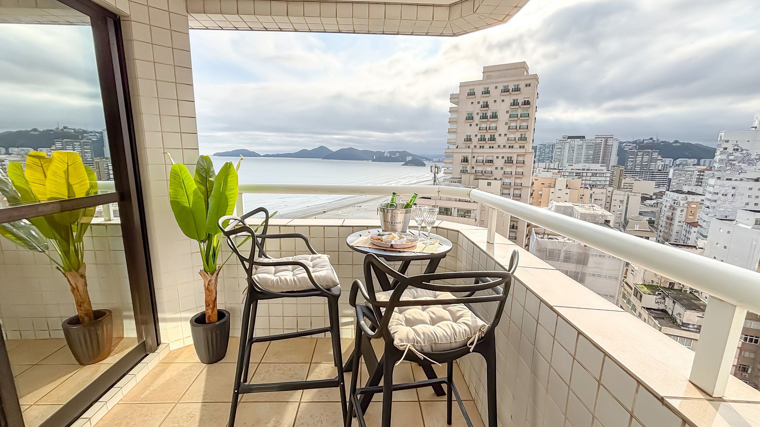 Un balcón con mesa y sillas, una planta en maceta y vistas a la playa y al paisaje urbano.