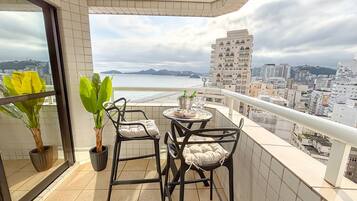 Un balcón con mesa y sillas, una planta en maceta y vistas a la playa y al paisaje urbano.