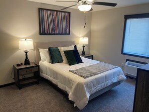Ein Schlafzimmer mit einem Bett, zwei Nachttischen mit Lampen, einem Deckenventilator, einem Fenster mit Jalousien und einem Wandbild.