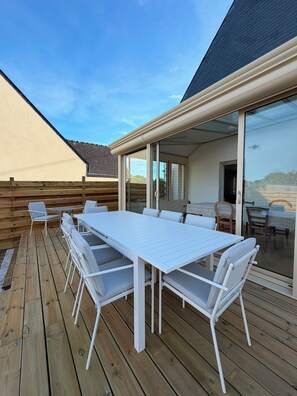 A covered outdoor patio with a white table and chairs, wooden deck flooring, and a glass door leading to an interior space. - L’Oasis – Family home for 12, enclosed garden, walk to beach (Saint-Pierre-Quiberon)