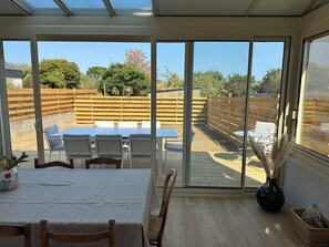 A sunlit patio with a table and chairs, a sliding glass door leading to a fenced backyard, and a potted plant on the table. - L’Oasis – Family home for 12, enclosed garden, walk to beach (Saint-Pierre-Quiberon)