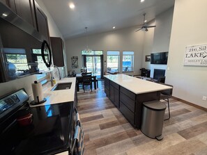 A modern kitchen with a large island, dark cabinetry, and a dining area with a view of the outdoors. - Seeley Lake Mountain Modern (Seeley Lake)