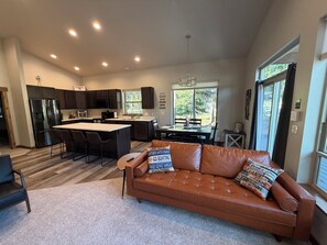 A modern kitchen with dark cabinets, a central island, and a dining area with a view of the outdoors. - Seeley Lake Mountain Modern (Seeley Lake)