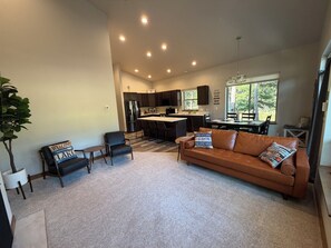A modern kitchen with dark cabinetry, a central island, and a dining area with a brown leather couch and chairs. - Seeley Lake Mountain Modern (Seeley Lake)