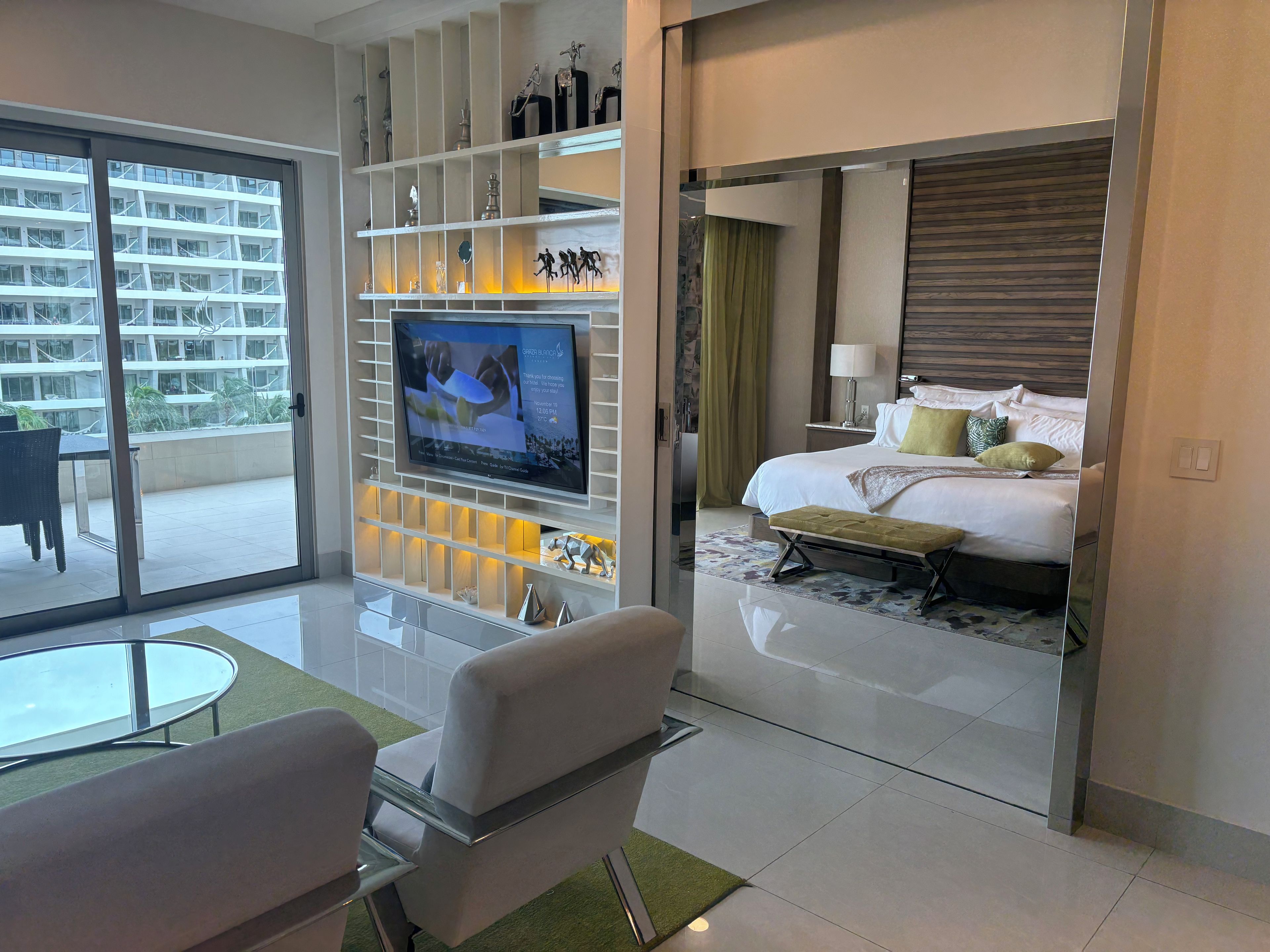 Una camera d'albergo con un letto, una TV, un'area salotto con tavolo e sedie, e un balcone con vista su un edificio.