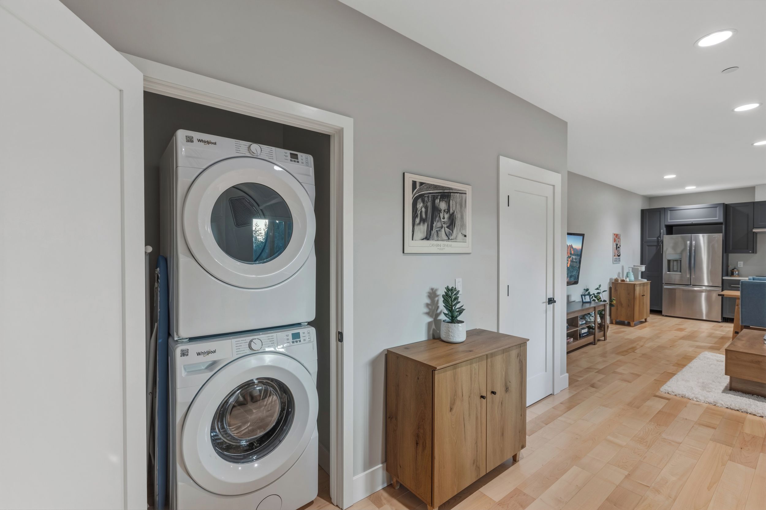 Une buanderie équipée de deux lave-linge Whirlpool superposés, d’un meuble en bois et d’une porte donnant sur une cuisine avec un réfrigérateur en acier inoxydable.
