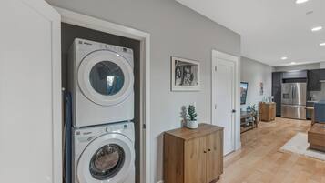Une buanderie équipée de deux lave-linge Whirlpool superposés, d’un meuble en bois et d’une porte donnant sur une cuisine avec un réfrigérateur en acier inoxydable.