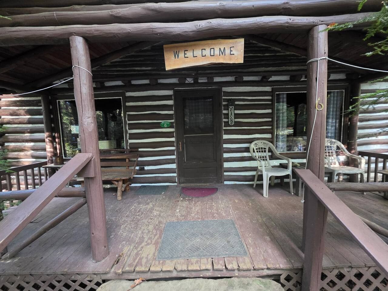 A wooden cabin with a 'WELCOME' sign, a porch with a ramp, and plastic chairs.