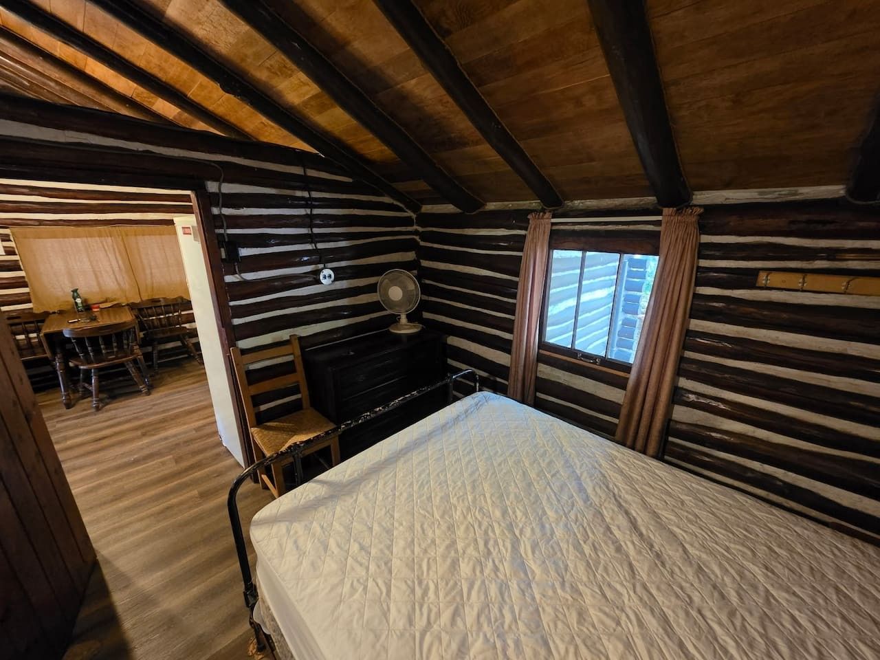 A cabin-style room with a bed, a desk, and a chair. There is a window with curtains and a fan on the wall.