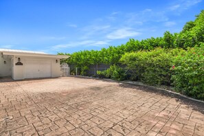A paved driveway leading to a white garage with a number sign, surrounded by lush green foliage and a clear blue sky. - Canal-View Terrace, Family-Friendly, 5 Min to MIA (Miami Springs)