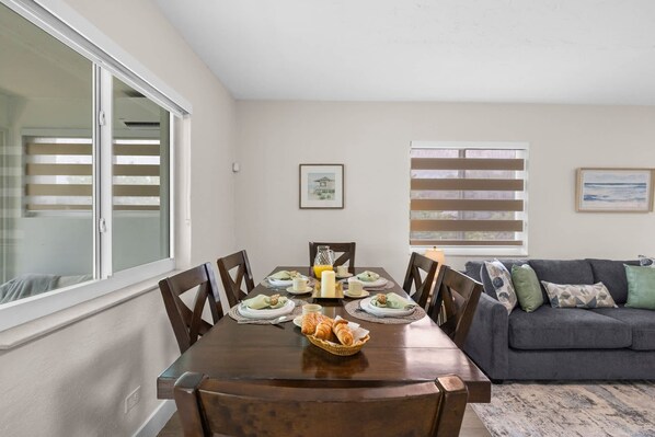 A dining area with a wooden table set for four, surrounded by chairs. There is a window with blinds on the left, a framed picture on the wall, and a grey sofa with cushions in the background. - Canal-View Terrace, Family-Friendly, 5 Min to MIA (Miami Springs)