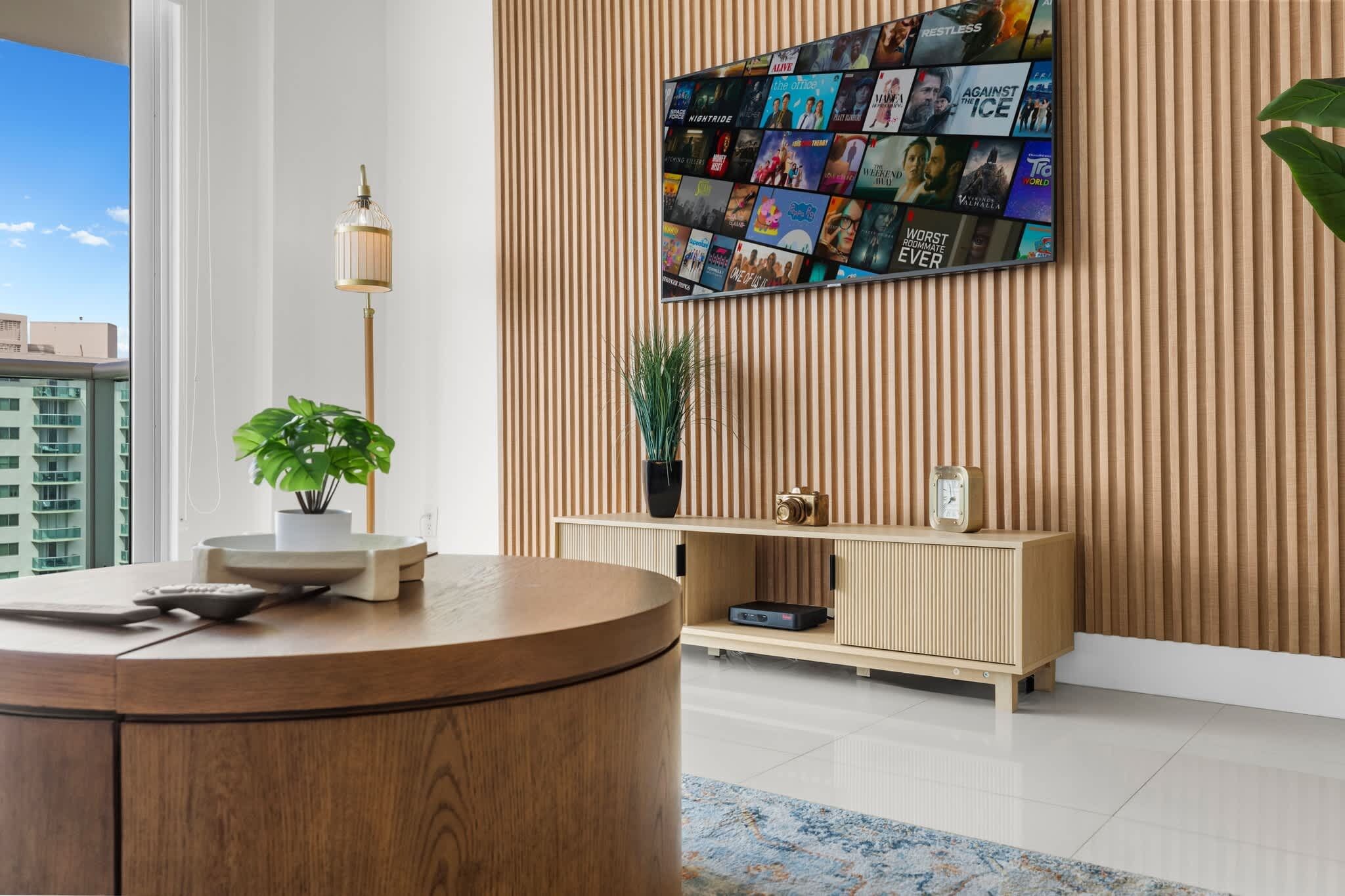 A modern lobby with a wooden reception desk, a wall-mounted TV displaying movie posters, a potted plant, and a vintage clock.