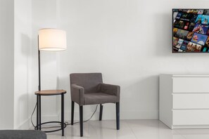 Ein modernes Zimmer mit grauem Sessel, kleinem runden Tisch, Stehlampe und einem an der Wand montierten Fernseher, der verschiedene Filmplakate zeigt.